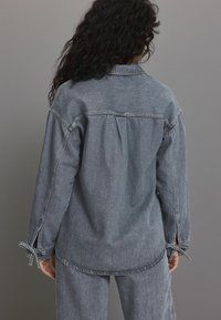 Graue Denimhemden mit einem lockeren Schnitt, seitlichem Bindebanddetail an den Ärmeln und einem Rückenpasse. Strukturiertes Gewebe mit subtilen Variationen.