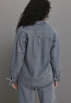 Chemise en denim gris avec une coupe décontractée, détail de lien sur les manches et un empiècement au dos. Tissu texturé avec des variations subtiles.