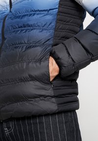 Gevoerde jas met een gradient ontwerp in blauwe en zwarte secties. Het materiaal lijkt glanzend met zichtbare stiksels en een veilige zak.