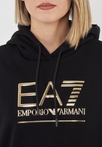Felpa nera con un grande logo dorato "EA7 EMPORIO ARMANI" sulla parte anteriore. Presenta cappucci con cordini e un tessuto soffice e texture.