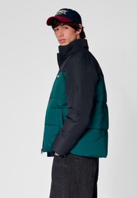 Kurtka puchowa w ciemnozielonym i granatowym kolorze, z wysokim kołnierzem, zamkiem błyskawicznym i pikowaną fakturą. Połączona z ciemnymi dżinsami i czapką.