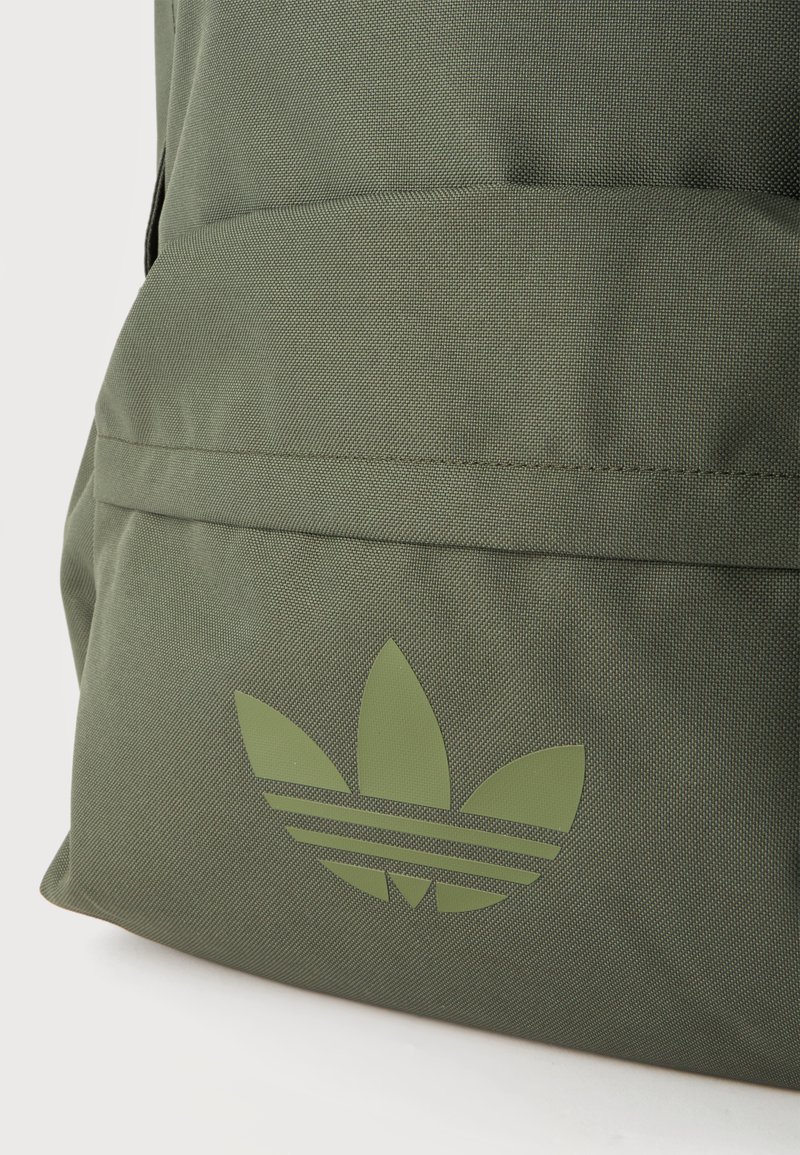 Gros plan sur un sac à dos vert olive avec un grand logo trèfle Adidas vert clair sur la poche avant.