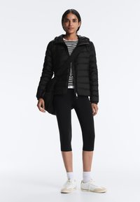 Nő fekete pehelydzsekiben, fekete rövidített leggingsben, csíkos ingben, fehér tornacipőben, fekete oldaltáskával a vállán, fehér háttér előtt áll.