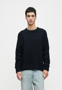 Mlad moški, oblečen v temno modro pulover s pletenim vzorcem in svetlo modre kavbojke, stoji pred enobarvno belo ozadjem.