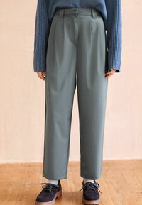 Pantaloni larghi verde chiaro con pieghe e finitura liscia, abbinati a scarpe scure e calzini chiari, su uno sfondo di pavimento in legno.