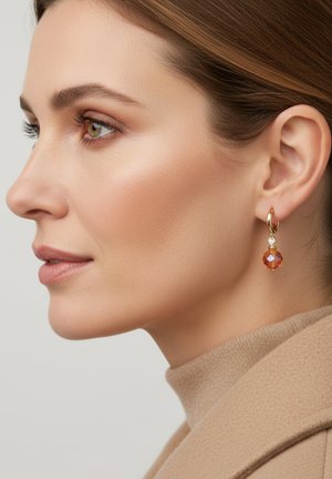 Gouden oorringen met een oranje kristalhangertje, tegen een neutrale achtergrond. Het onderwerp draagt een beige trui met hoge hals.