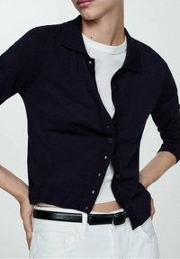 Námořnický svetr s límečkem, zapínáním na knoflíky a žebrovanými manžetami, nošený přes bílou košili a doplněný bílými kalhotami a černým páskem.