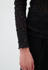 Černý top s dlouhým rukávem, který má složitý texturovaný krajkový design se scalloped lemem, spárován s hladkým černým materiálem pod ním.