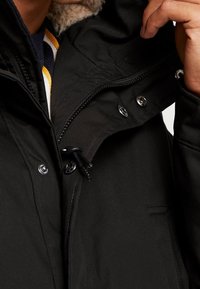 Parka negra con un exterior texturizado, que cuenta con un cierre de cremallera y botones a presión, capucha ajustable con cordón y un suave cuello forrado.