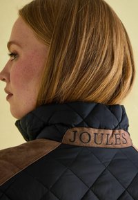 Veste matelassée noire avec épaule et col en daim marron, portant le mot « JOULES » brodé sur le col.