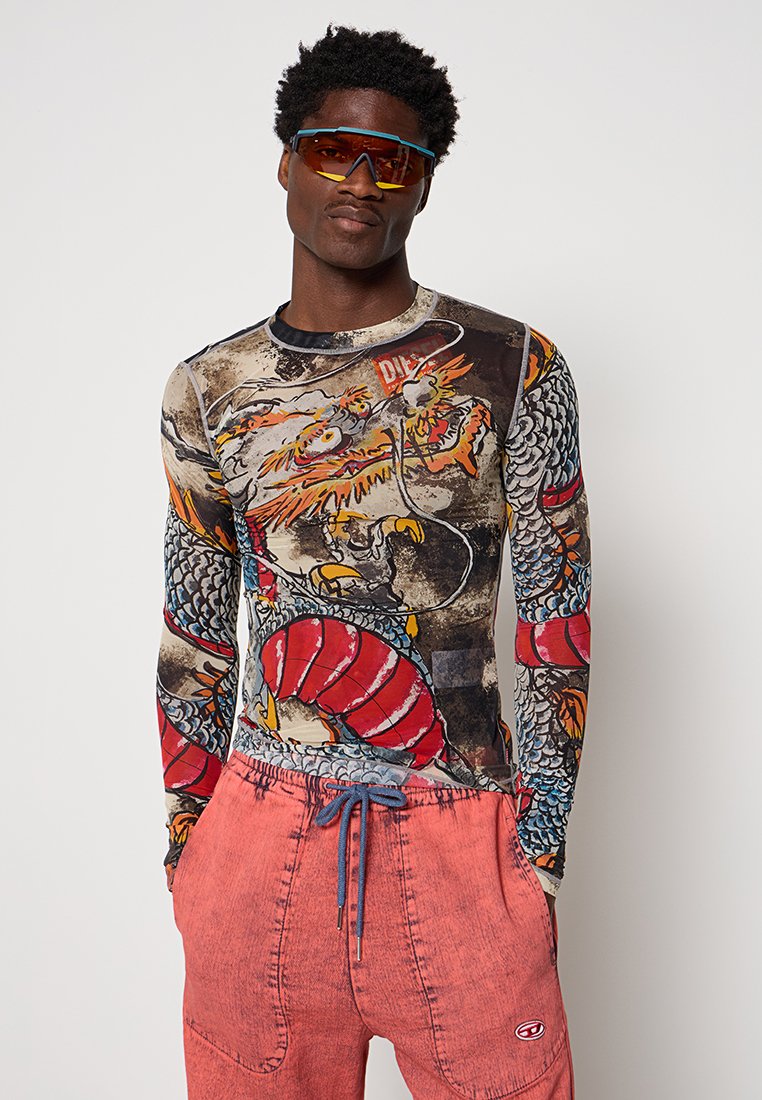 Langärmliges Shirt mit einem strukturierten Grafikdruck, der einen Drachen und einen Fisch zeigt. Kombiniert mit lockeren rosa Shorts und sportlichen Sonnenbrillen.