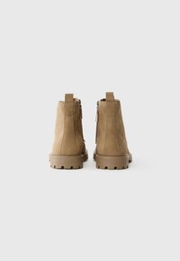 Par de botas de tobillo de ante color marrón claro con tiradores y cremalleras laterales, mostradas desde atrás sobre un fondo blanco liso.