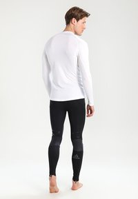 Ilga piedurkne balta sporta krekls, kas kombinēts ar melnajām legingām ar logotipu, rāvējslēdzēja potītēm un teksturētām akcentiem uz kājām.