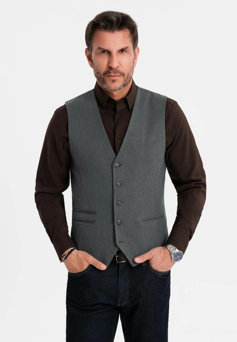 Mann trägt ein dunkelbraunes Hemd, eine graue, geknöpfte Weste, dunkle Jeans, eine silberne Armbanduhr und hat die Hände in den Taschen vor einem schlichten weißen Hintergrund.