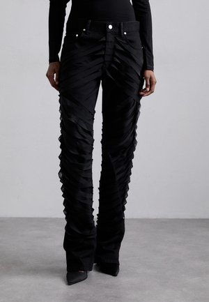 Pantalons en denim noir avec un design superposé comportant des bandes de tissu horizontales, taille haute et bas évasés. Accents en métal argenté visibles.