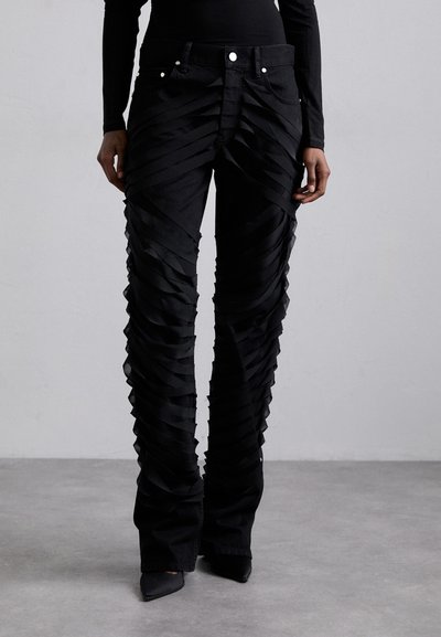 Pantalons en denim noir avec un design superposé comportant des bandes de tissu horizontales, taille haute et bas évasés. Accents en métal argenté visibles.