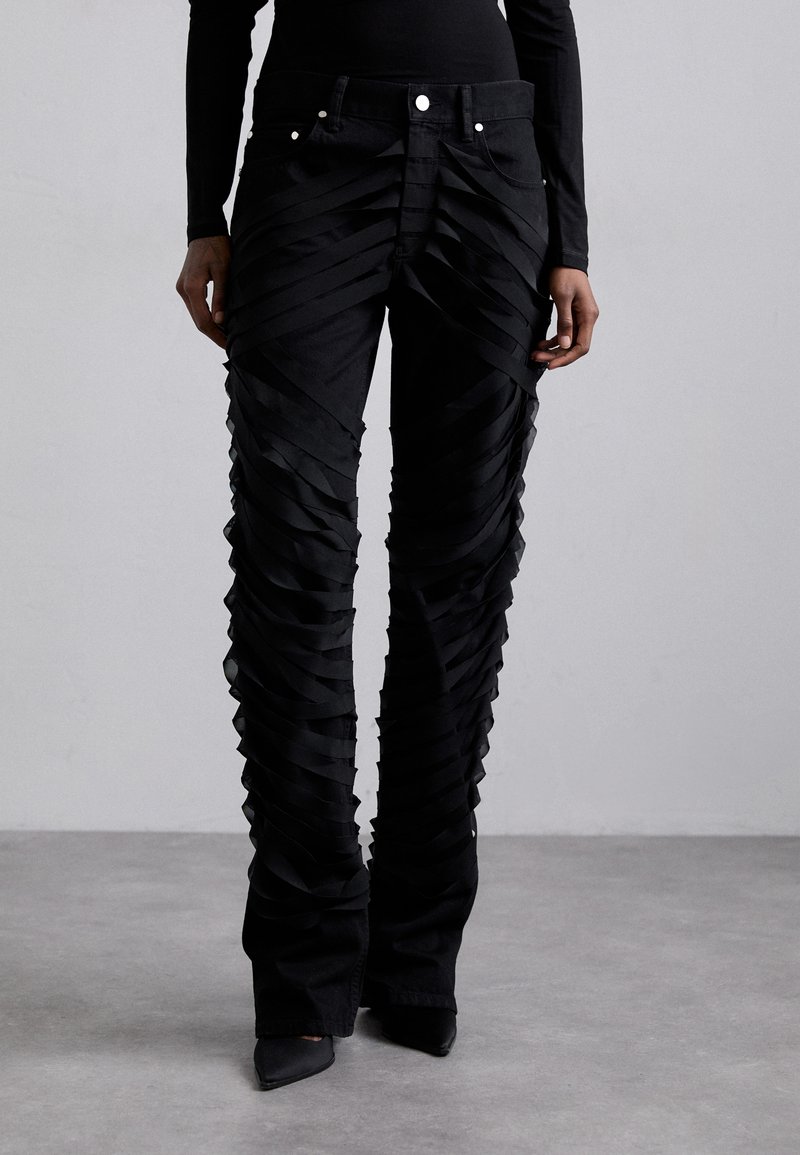 Pantalons en denim noir avec un design superposé comportant des bandes de tissu horizontales, taille haute et bas évasés. Accents en métal argenté visibles.