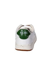 Zapatilla blanca con parte superior de cuero texturizado, parche verde en el talón y suela salpicada de marrón. Diseño minimalista con detalle de collar acolchado.