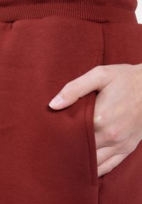 Bordowe dresy o miękkiej fakturze, z prążkowanym pasem i boczną kieszenią, z rękami częściowo włożonymi do kieszeni.