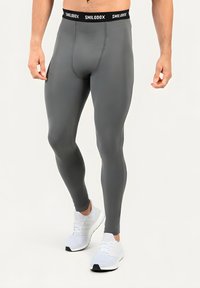Homme portant des leggings de compression gris foncé et des chaussures de sport blanches, debout devant un fond clair.