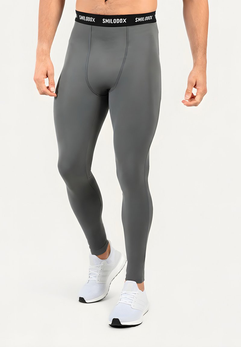 Homme portant des leggings de compression gris foncé et des chaussures de sport blanches, debout devant un fond clair.