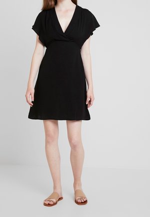 Kvinde iført en kort sort V-hals slå-om-kjole og beige flade sandaletter med åbne tæer, stående mod en ensartet lys baggrund.