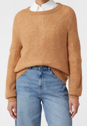 Femme portant un pull en tricot couleur camel avec des sequins subtils par-dessus une chemise blanche à col, associée à un jean taille haute bleu clair.