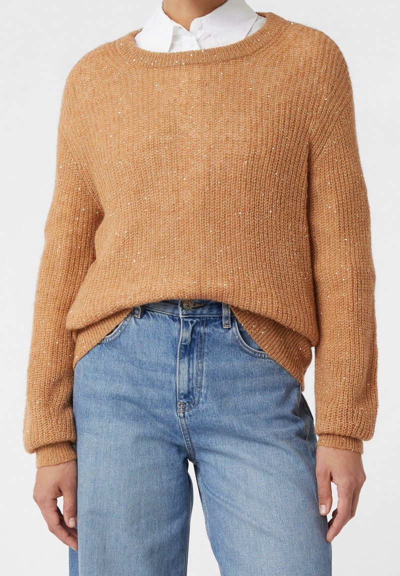 Femme portant un pull en tricot couleur camel avec des sequins subtils par-dessus une chemise blanche à col, associée à un jean taille haute bleu clair.