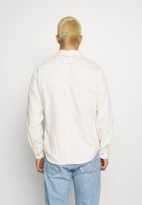 Camicia button-up beige realizzata in cotone, con maniche lunghe, colletto e un'etichetta quadrata sul retro con cucitura rossa.