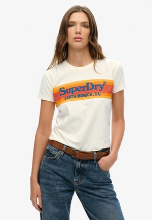 Jonge vrouw die een wit SuperDry T-shirt draagt met oranje en gele strepen en blauwe tekst, gecombineerd met een blauwe spijkerbroek en een bruine riem.