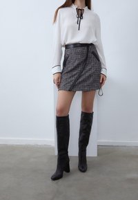 Bílá halenka s černou stuhou a manžetami, spárovaná s černobílou kostkovanou sukní s postranní kapesní a koženými doplňky. Černé kozačky po kolena.