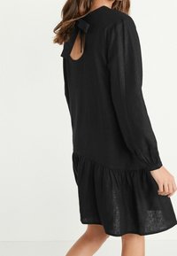 Mujer vestida con un vestido negro de manga larga con falda recogida y un corte en la espalda atado con un lazo en el escote.
