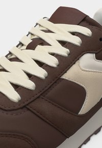 Baskets marron et beige avec lacets blancs, gros plan montrant la texture du tissu et le détail des lacets sur le côté et la zone de la languette.