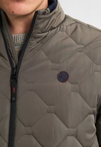 Man in een olijfgroene gewatteerde jas met zwarte rits, hoge kraag en een rond logo op de borst, over een lichtgrijze gebreide trui.