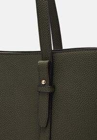 Sac fourre-tout en cuir vert texturé avec une seule sangle, présentant un détail de bouton-pression doré et un bord noir contrastant le long des bords.