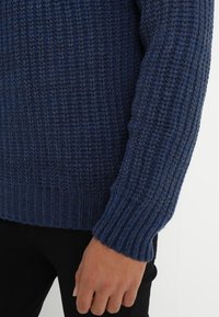Sweter dzianinowy o prążkowanej fakturze, w głębokim granatowym kolorze. Posiada długie rękawy oraz dopasowany dół. Materiał wydaje się miękki i ciepły.