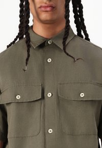 Chemise verte à manches courtes avec un col boutonné, deux poches poitrine et des boutons de couleur claire. Le tissu semble texturé et léger.