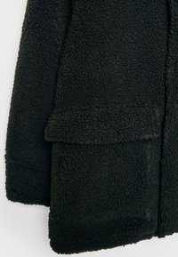 Svart fleecemantel med texturerad yta, som har en framficka och öppna kanter. Tyget ser mjukt och varmt ut, vilket gör det lämpligt för kallt väder.