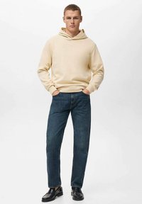 Beige hættejakke med justerbar snøre, parret med mørkeblå jeans. Outfittet afsluttes med sorte sko. Simpel, afslappet pasform.