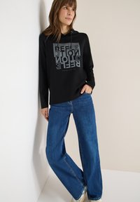 Černá mikina s potiskem textu v šedé barvě a modré široké džíny. Mikina má šňůrku a volný střih.