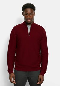 Burgunderfarbener Pullover mit halber Reißverschluss-Kragen, gerippter Textur und einfarbigem Design. Verfügt über lange Ärmel und einen figurbetonten Bund. Wird über einem grauen Shirt getragen.