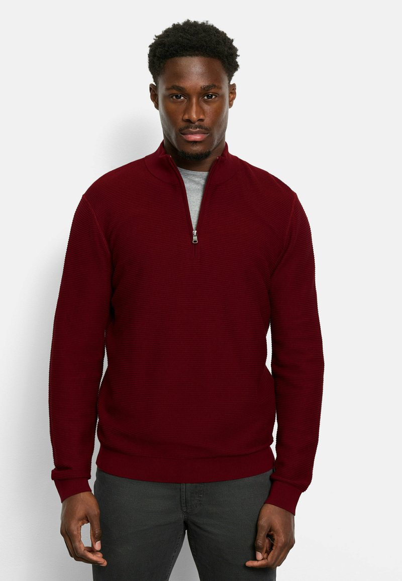 Burgunderfarbener Pullover mit halber Reißverschluss-Kragen, gerippter Textur und einfarbigem Design. Verfügt über lange Ärmel und einen figurbetonten Bund. Wird über einem grauen Shirt getragen.