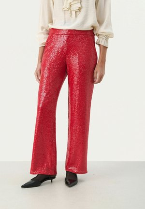 Pantalones de pata ancha rojos con lentejuelas, combinados con una blusa cremosa con cuello con volantes y zapatos negros puntiagudos de tacón bajo sobre un suelo claro.