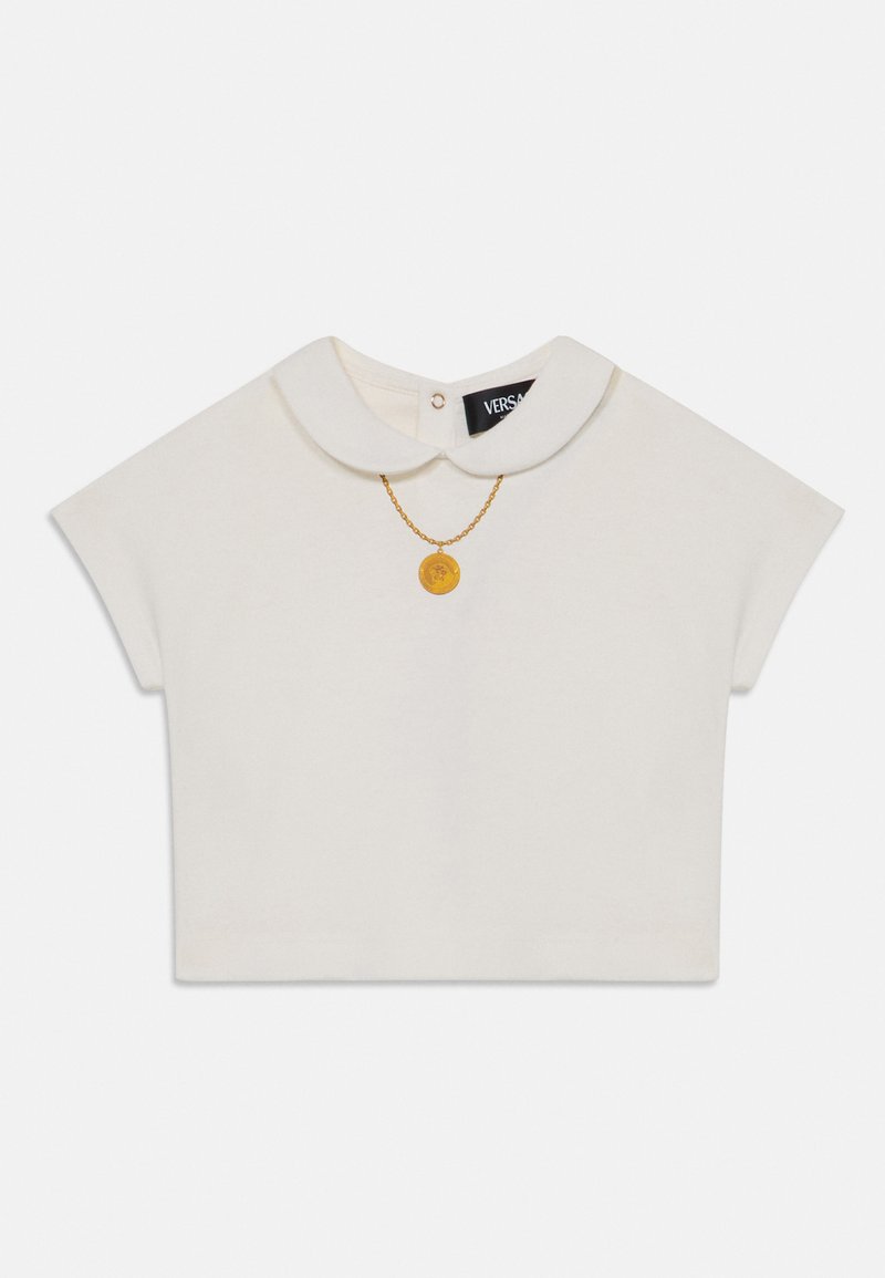 Λευκή κοντομάνικη μπλούζα με γιακά Peter Pan, διαθέτοντας μια χρυσή αλυσίδα με κυκλική κρέμαση και μια μαύρη ετικέτα στον λαιμό.