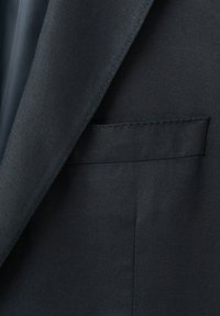 Nærbillede af mørk skræddersyet jakkesætjakke forfra, der viser revers og syningsdetaljer ved paspoleret lomme.