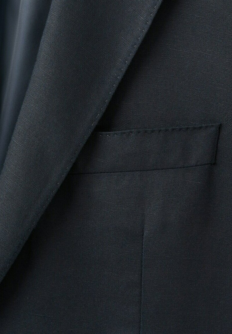 Gros plan sur l'avant d'une veste de costume sombre sur mesure montrant les détails des coutures du revers et de la poche passepoilée.