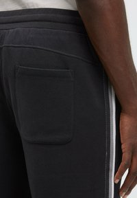 Schwarze Jogginghose mit seitlichem grauen Streifen, elastischem Bund und einer aufgesetzten Tasche auf der Rückseite. Hergestellt aus weichem, strukturiertem Stoff.