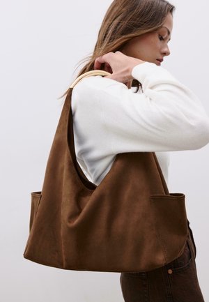 Žena u bijelom džemperu drži veliku smeđu torbu od brušene kože s okruglim smeđim ručkama preko ramena na neutralnoj pozadini.