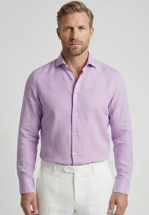 Homme blond avec barbe portant une chemise lilas clair à boutons et un pantalon blanc, debout devant un fond clair uni.