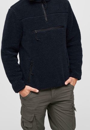 Homme portant un pull en polaire noir avec poches zippées sur la poitrine et les côtés, associé à un pantalon cargo vert olive, mains dans les poches.